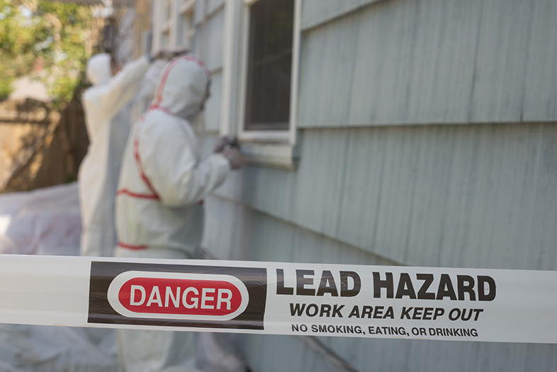 Two people in hazmat suits in an area behind lead hazard warning tape