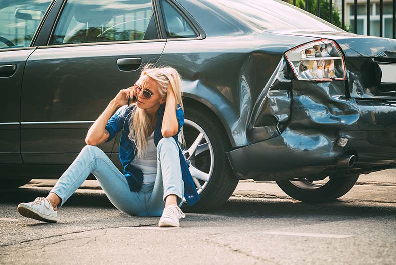 Distressed Minnesota woman sitting and talking on the phone beside her damaged car after an accident