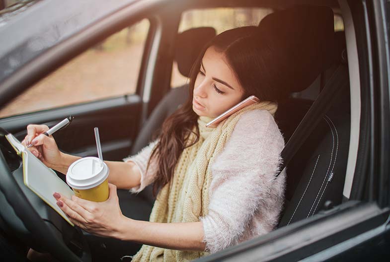 Young Wisconsin woman talking on her phone, writing in a notebook, and drinking coffee while driving