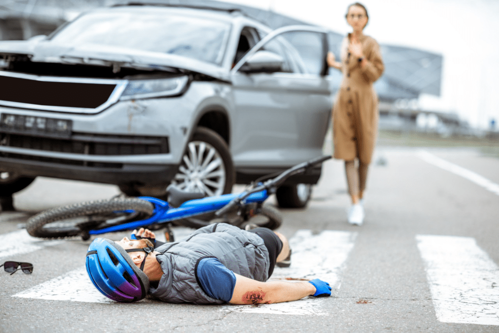 An injured cyclist lies on the ground next to a wrecked vehicle while a concerned woman examines the scene of the accident. 