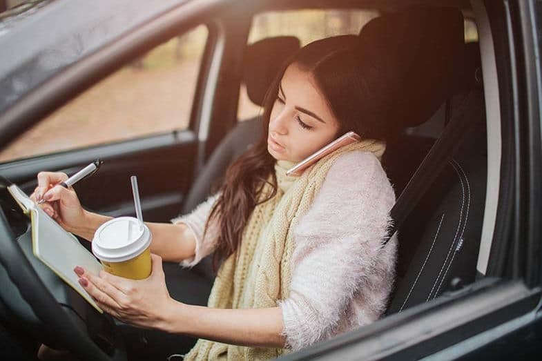 Young Wisconsin woman talking on her phone, writing in a notebook, and drinking coffee while driving