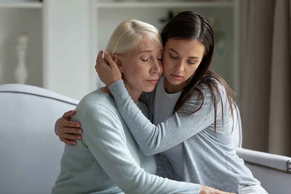 Young woman consoling and comforting her distressed middle-aged mother on a cozy home couch. Compassionate support and love between generations.