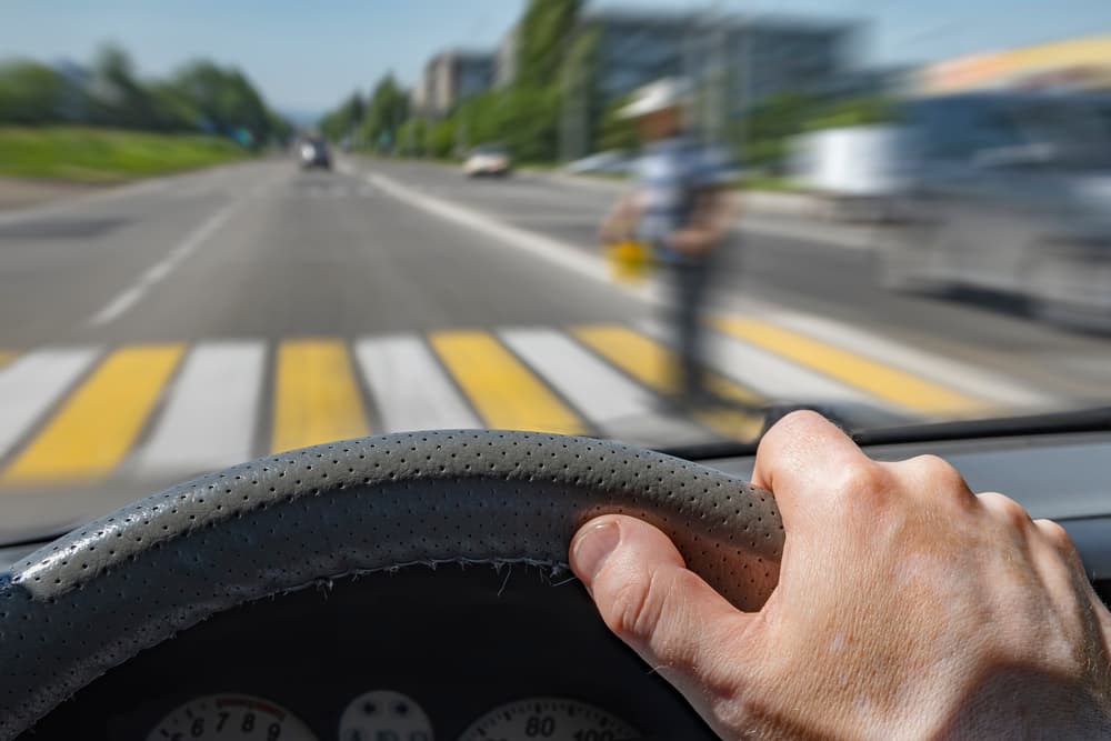 Driver's hand on a fast-moving car steering wheel near a pedestrian crossing, emphasizing the risk of potential collision.