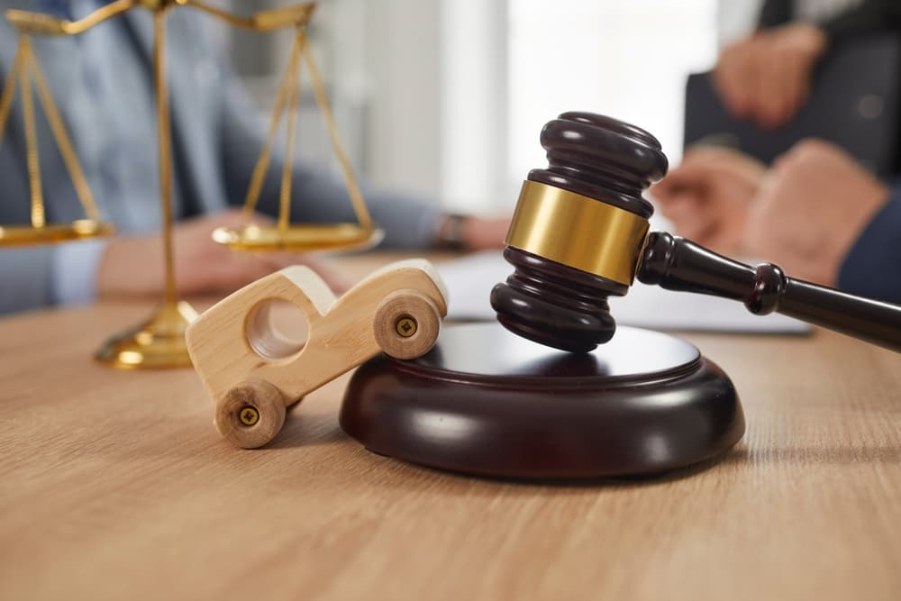 Small wooden toy car, sound block, gavel, and scales of justice on an office table, with people discussing in the background, representing legal services and accident law.