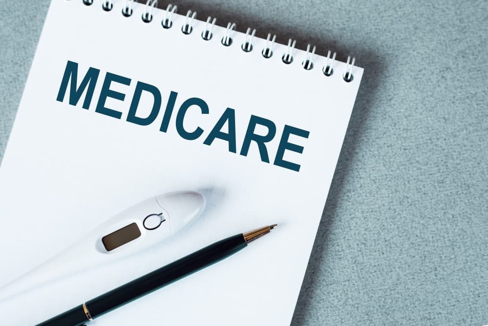 Notepad with the word 'MEDICARE' written on it, placed on a blue surface next to a black pen and an electronic thermometer, representing a medical theme.