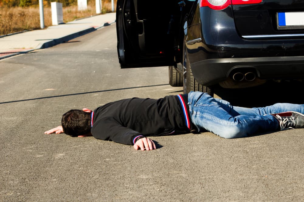 A man lying on the road behind a black car after being hit in a pedestrian collision.