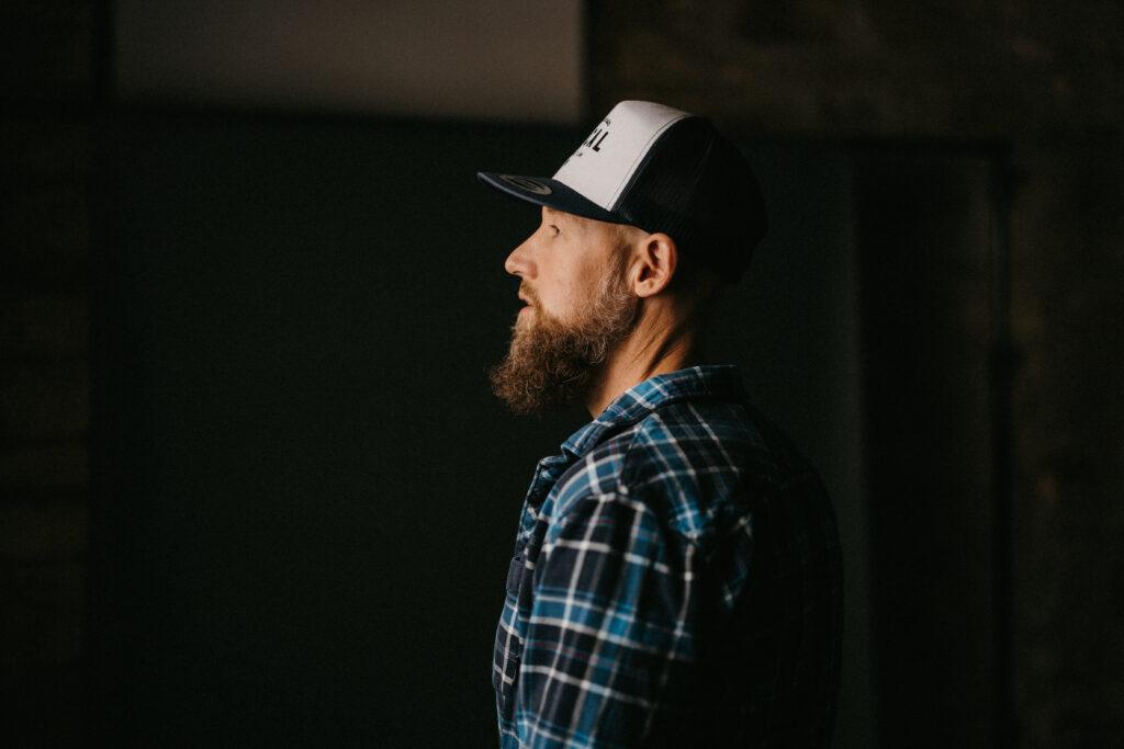 Bearded man wearing a baseball cap and plaid shirt, shown in side profile against a dark, moody background.