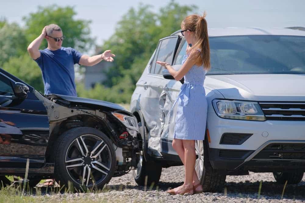 Two drivers standing beside their damaged vehicles after a car accident, discussing fault near a rural roadside.