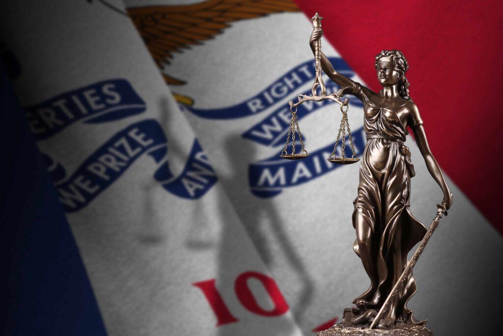 Lady Justice statue holding scales in front of the Iowa state flag, representing Iowa courts and state legal system.