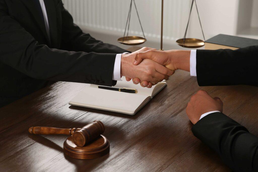 Scales of justice balancing cash and judge’s gavel in law office. Attorney and client shaking hands over desk with gavel and scales of justice, symbolizing legal agreement, consultation, or case settlement.