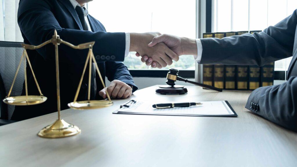 Two attorneys shaking hands across desk with scales of justice and gavel, symbolizing legal agreement, settlement, or successful case resolution. Two attorneys shaking hands across desk with scales of justice and gavel, symbolizing legal agreement, settlement, or successful case resolution.